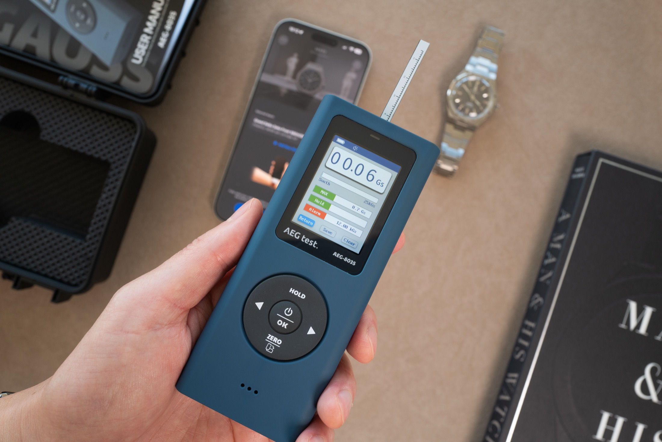 Hand holding AEGTEST 8035 gaussmeter with telescopic probe extended, display showing 0.06 Gauss